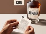 Nahaufnahme einer Hand, die ein Glas Aviation Gin hält, mit einem Verkostungsnotizblatt und einer unscharfen Flasche im Hintergrund; modernes Gemälde mit braunem Farbschema - Aviation Gin Test.